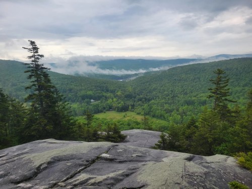 A scenic view from a rocky outcrop, overlooking lush green valleys and misty mountains under a cloudy sky.