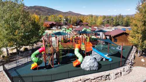 Colorful playground with slides, climbing structures, and a rock feature, surrounded by trees and mountains in the background.