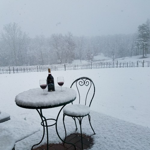 A snowy landscape with a table set for two, featuring a bottle of wine and two glasses, surrounded by a vineyard.