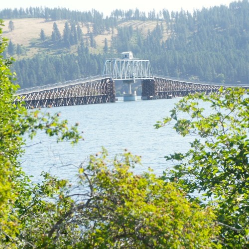 A bridge spans a calm body of water, surrounded by green trees and rolling hills in the background.