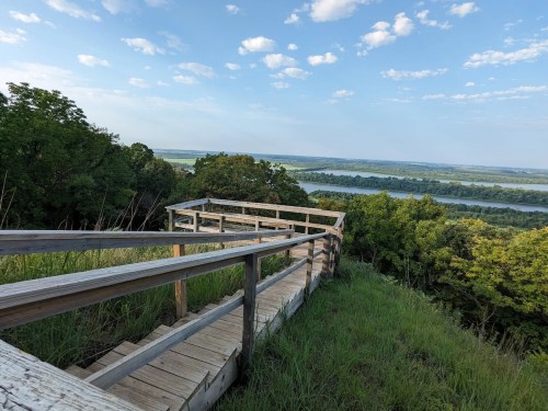 A wooden walkway leads to a scenic overlook of a river and lush green landscape under a blue sky with clouds.