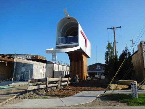A large, modern mailbox structure stands on a wooden base, surrounded by construction and utility poles under a clear blue sky.