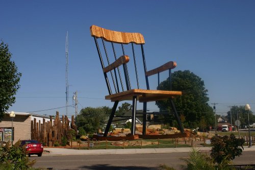 A giant wooden rocking chair stands in a park, surrounded by trees and a clear blue sky.
