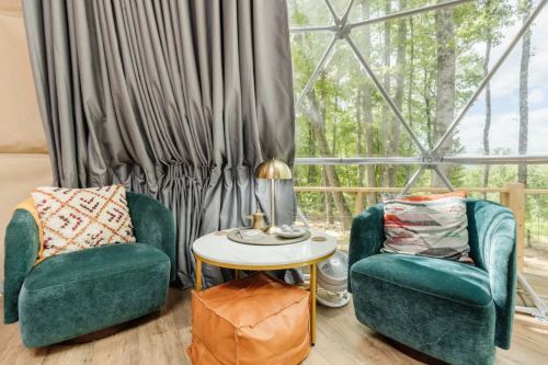 Cozy interior with two green chairs, a round table, and a warm orange pouf, surrounded by large windows and nature.