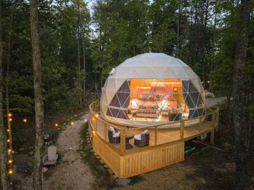 A geodesic dome cabin nestled in a forest, featuring a wooden deck and surrounded by trees and outdoor seating.