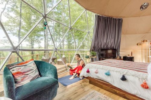 A cozy, modern geodesic dome interior with a bed, a green chair, and large windows overlooking trees. A woman reads by the window.