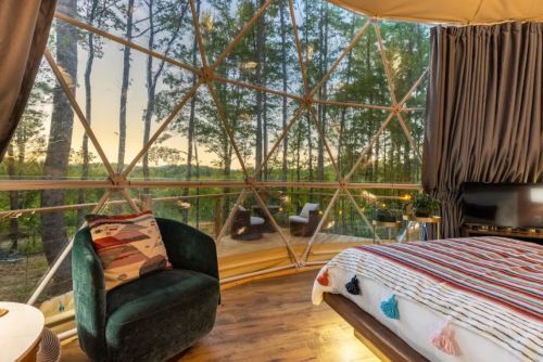 Cozy geodesic dome interior with a bed, chair, and large windows showcasing a forest view at sunset.
