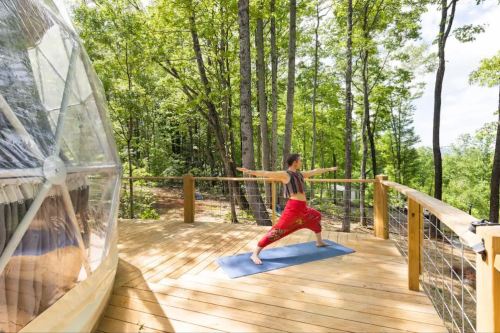 A person practicing yoga in warrior pose on a wooden deck surrounded by trees and a geodesic dome.