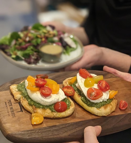 Two toasted bread slices topped with pesto, burrata, and colorful cherry tomatoes, served with a side salad.
