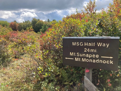 Signpost indicating the halfway point of the MSG trail, with directions to Mt. Sunapee and Mt. Monadnock, surrounded by autumn foliage.