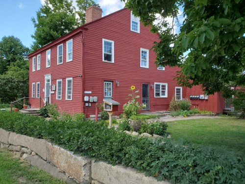 A red two-story house surrounded by greenery and a stone wall, with a garden and bird feeder in the front yard.