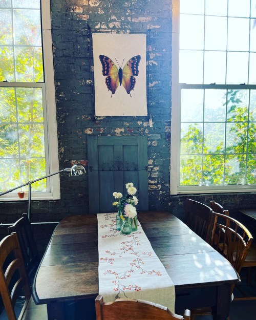 A cozy dining area with a wooden table, floral centerpiece, and a butterfly artwork on the wall, surrounded by large windows.