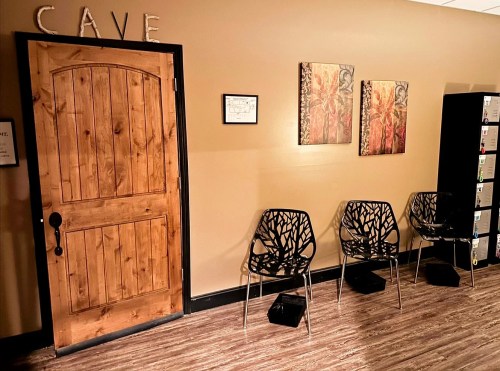 A wooden door labeled "CAVE" next to two decorative wall art pieces and three modern chairs in a cozy room.