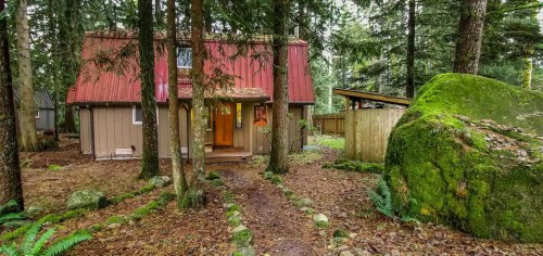 A cozy cabin with a red roof nestled among tall trees and mossy rocks, surrounded by a forested landscape.