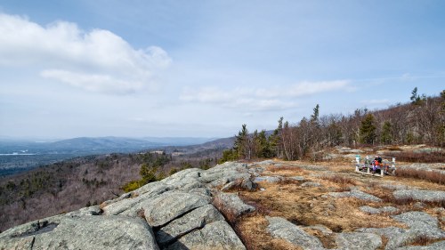 A scenic mountain view with rocky terrain, sparse trees, and a person sitting on a bench overlooking the landscape.