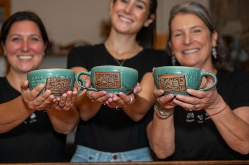 Three women hold matching green mugs with "Inspire Cafe" printed on them, smiling at the camera.