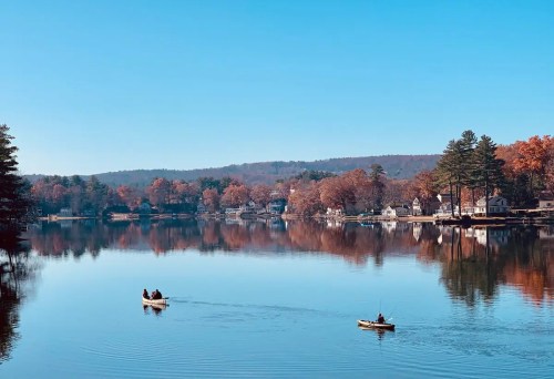 Two small boats on a calm lake surrounded by autumn trees and reflections of houses in the water under a clear blue sky.