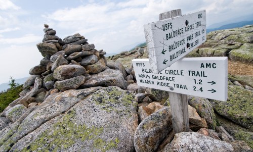 A rocky summit with a stacked stone cairn and directional signs for hiking trails in a mountainous area.