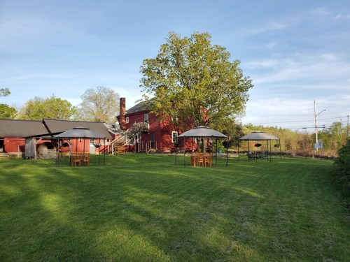 A red barn-style house with outdoor seating under canopies on a grassy lawn, surrounded by trees and blue sky.