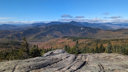A panoramic view of mountains and colorful autumn foliage under a clear blue sky. Rocky foreground with trees.