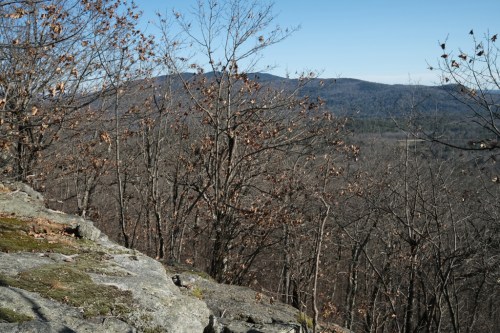 A rocky outcrop overlooks a forested valley with bare trees and distant mountains under a clear blue sky.
