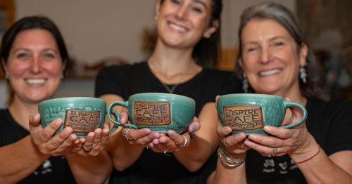 Three women hold matching green mugs with "Inspire Café" engraved, smiling at the camera in a cozy café setting.