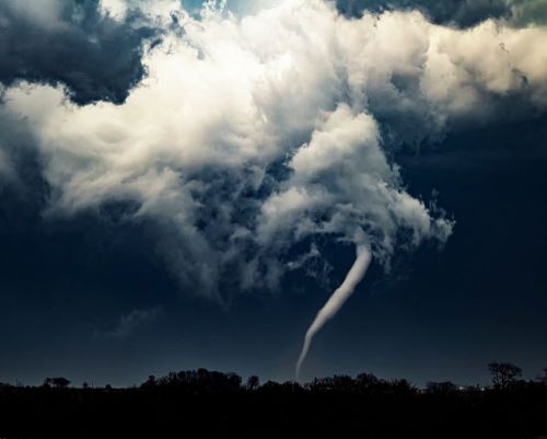 A dark sky with swirling clouds and a visible tornado touching down near a tree line.