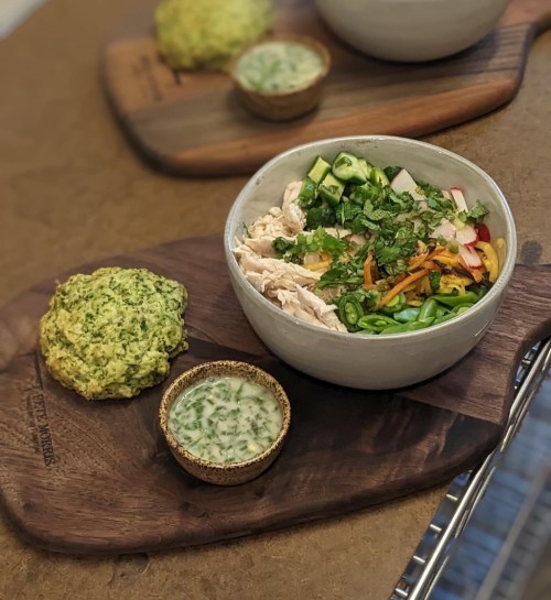 A bowl of salad with chicken, vegetables, and herbs, served with a green scone and a small bowl of dressing.