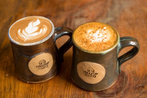 Two mugs of coffee with latte art, one light and creamy, the other spiced, on a wooden table.