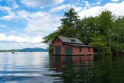 A red boathouse on a calm lake, surrounded by trees and mountains under a partly cloudy sky.
