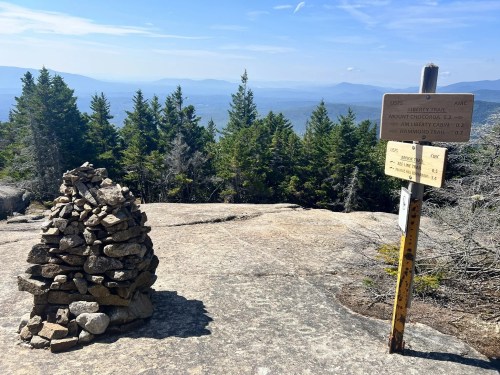 A stone cairn on a rocky mountain summit with a trail sign and scenic views of distant mountains and trees.