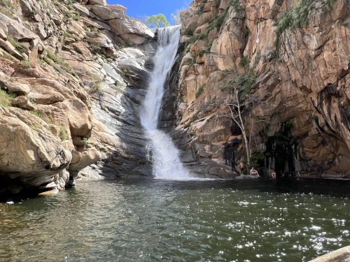 A scenic waterfall cascading down rocky cliffs into a serene pool, surrounded by lush greenery and sunlight.