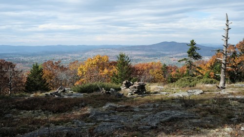 A scenic view of autumn foliage with mountains in the background and rocky terrain in the foreground.