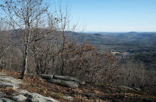 A scenic view from a rocky outcrop, showcasing rolling hills and a distant lake under a clear blue sky.