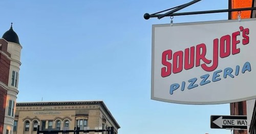 Sign for Sour Joe's Pizzeria hanging above a street, with buildings and a clear blue sky in the background.
