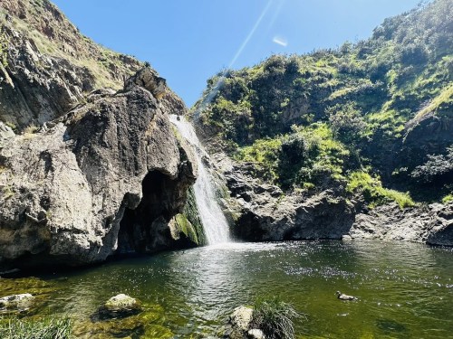 A serene waterfall cascading into a clear pool, surrounded by lush greenery and rocky cliffs under a bright blue sky.