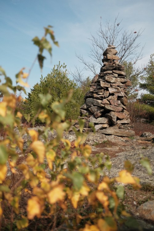 A stone cairn stands tall among greenery and autumn leaves under a clear blue sky.