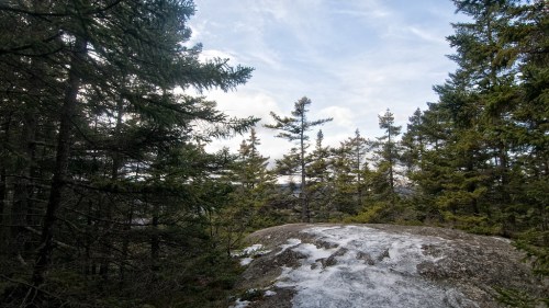 A rocky outcrop surrounded by evergreen trees under a cloudy sky, with patches of snow on the ground.