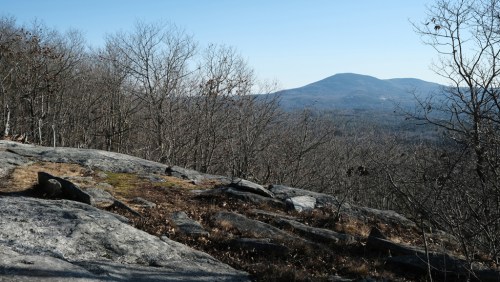 A rocky landscape with sparse trees, overlooking a distant mountain under a clear blue sky.