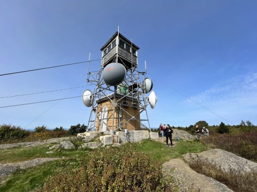 A tall communication tower with satellite dishes, surrounded by rocky terrain and people walking nearby under a clear blue sky.
