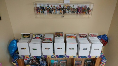 A collection of comic book boxes lined up, with action figures displayed above in a glass case.