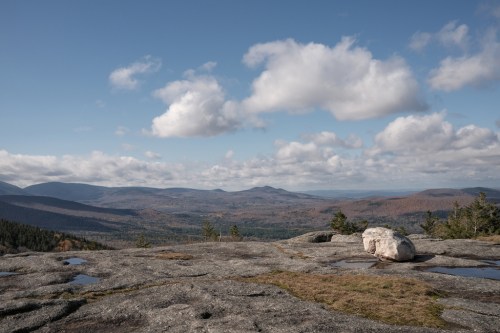 A rocky landscape with a large boulder, overlooking rolling hills and a blue sky with scattered clouds.