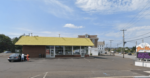 A fast-food restaurant with a yellow roof, parked cars, and a few people outside on a sunny day.