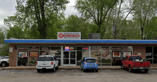 A small restaurant with a blue awning, featuring signs for donuts, gyros, and falafel, surrounded by trees.