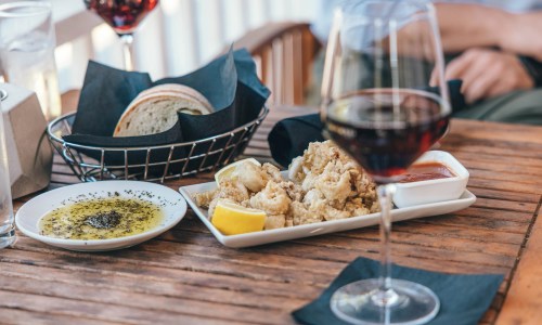 A table setting with a glass of red wine, fried calamari with lemon, dipping sauce, and a basket of bread.