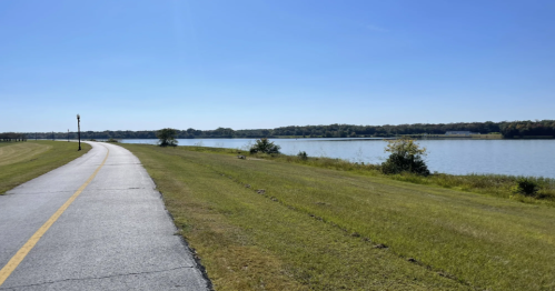 A paved path curves alongside a calm lake, with green grass and trees under a clear blue sky.