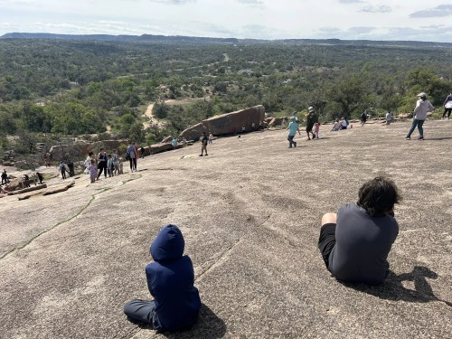 A child in a blue hoodie sits on a rocky slope, while others explore the area in the background under a cloudy sky.