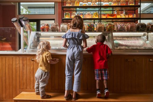 Three children stand at an ice cream counter, looking at various toppings and flavors in a bright, inviting shop.