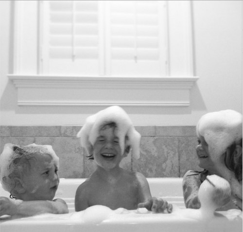 Three children play in a bathtub filled with bubbles, laughing and enjoying their bath time together.