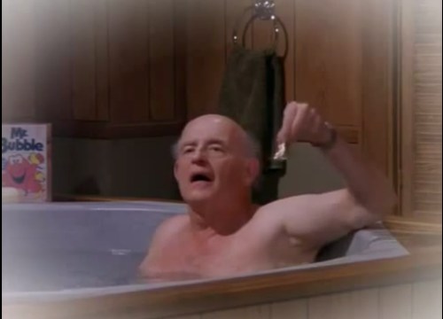An older man in a bathtub holds up a small item, with a towel hanging nearby and a bubble bath product in the background.